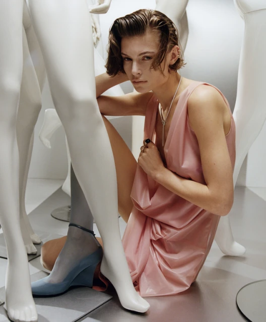 Fashionable woman poses amongst white mannequins, wearing a pale pink dress and blue high heels in a studio environment.
