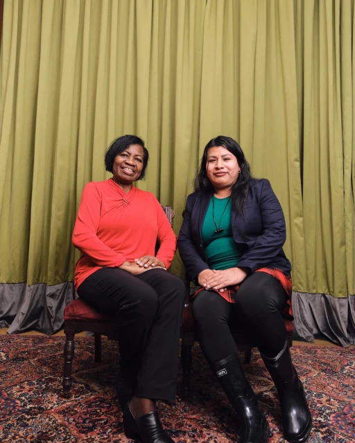 Two women, one in red and one in a blue jacket, sit side by side on antique chairs in front of a sage-green curtain indoors.
