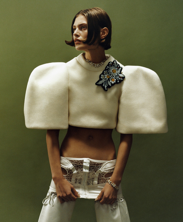 A female fashion model poses in a cropped, puffy-sleeved sweater and white pants against a green background in a studio shot.