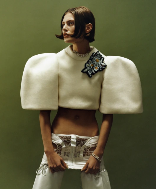 A female fashion model poses in a cropped, puffy-sleeved sweater and white pants against a green background in a studio shot.