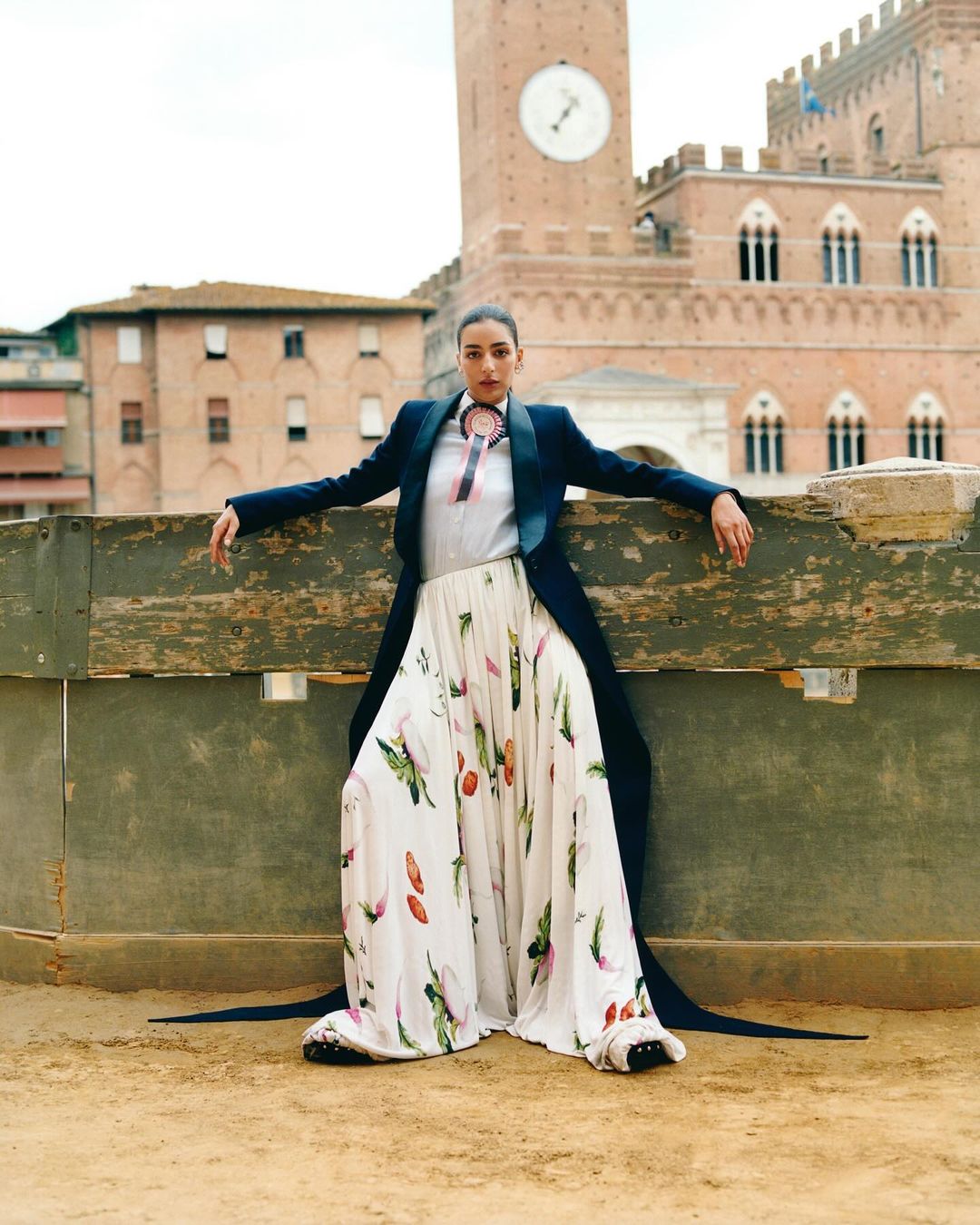 Adult female in a long printed skirt and blue blazer poses outdoors in a city square with a large clock tower in the background.