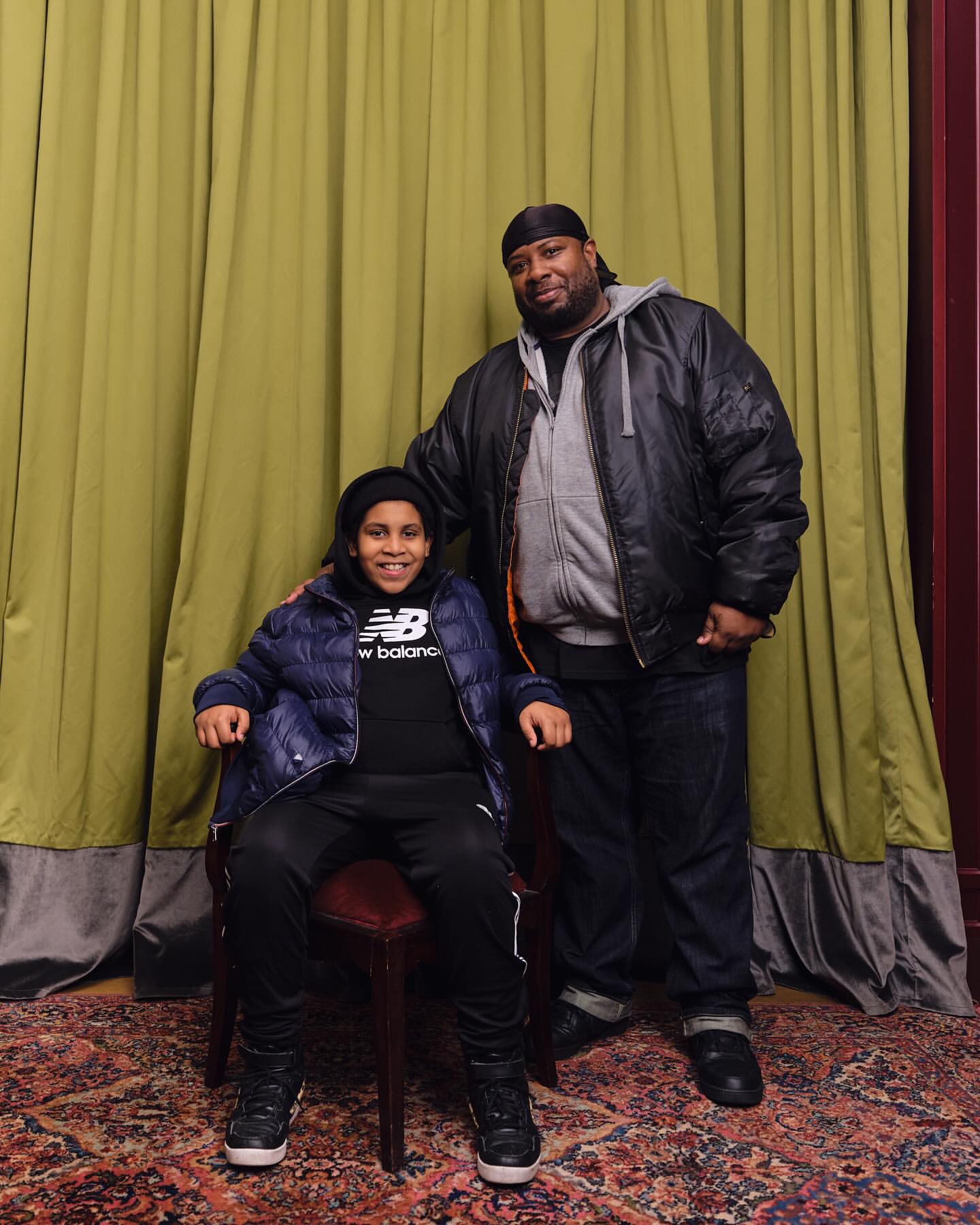 Portrait of an adult man in a jacket with a young person wearing a New Balance sweatshirt, both posed in front of a curtain.