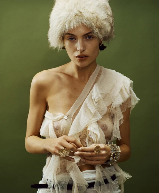 Portrait of a fair-skinned adult female in a sheer ruffled top and fur hat, posing against a green background in a fashion-style photo.