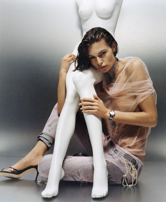 A young woman with wet hair poses beside a white mannequin in a fashion editorial shot in a studio setting with moody lighting and silver backdrop.