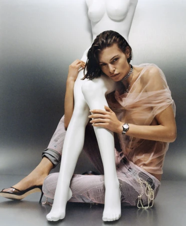 A young woman with wet hair poses beside a white mannequin in a fashion editorial shot in a studio setting with moody lighting and silver backdrop.