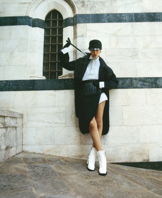 A fashionable adult female poses in front of a stone building wearing a black coat, white collared shirt, black skirt, hat, and white boots.