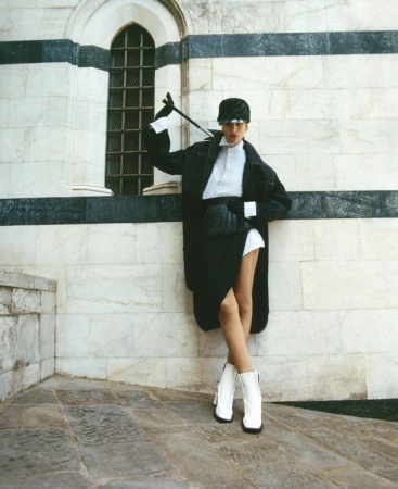 A fashionable adult female poses in front of a stone building wearing a black coat, white collared shirt, black skirt, hat, and white boots.