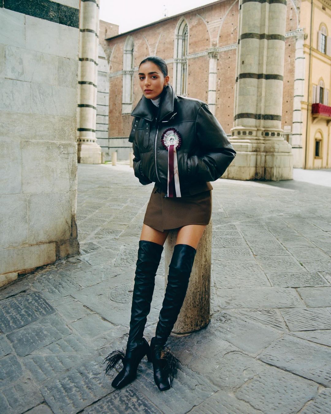 A woman in a dark leather puffer jacket and thigh-high boots posing outdoors in a cobblestone square framed by classical stone architecture.