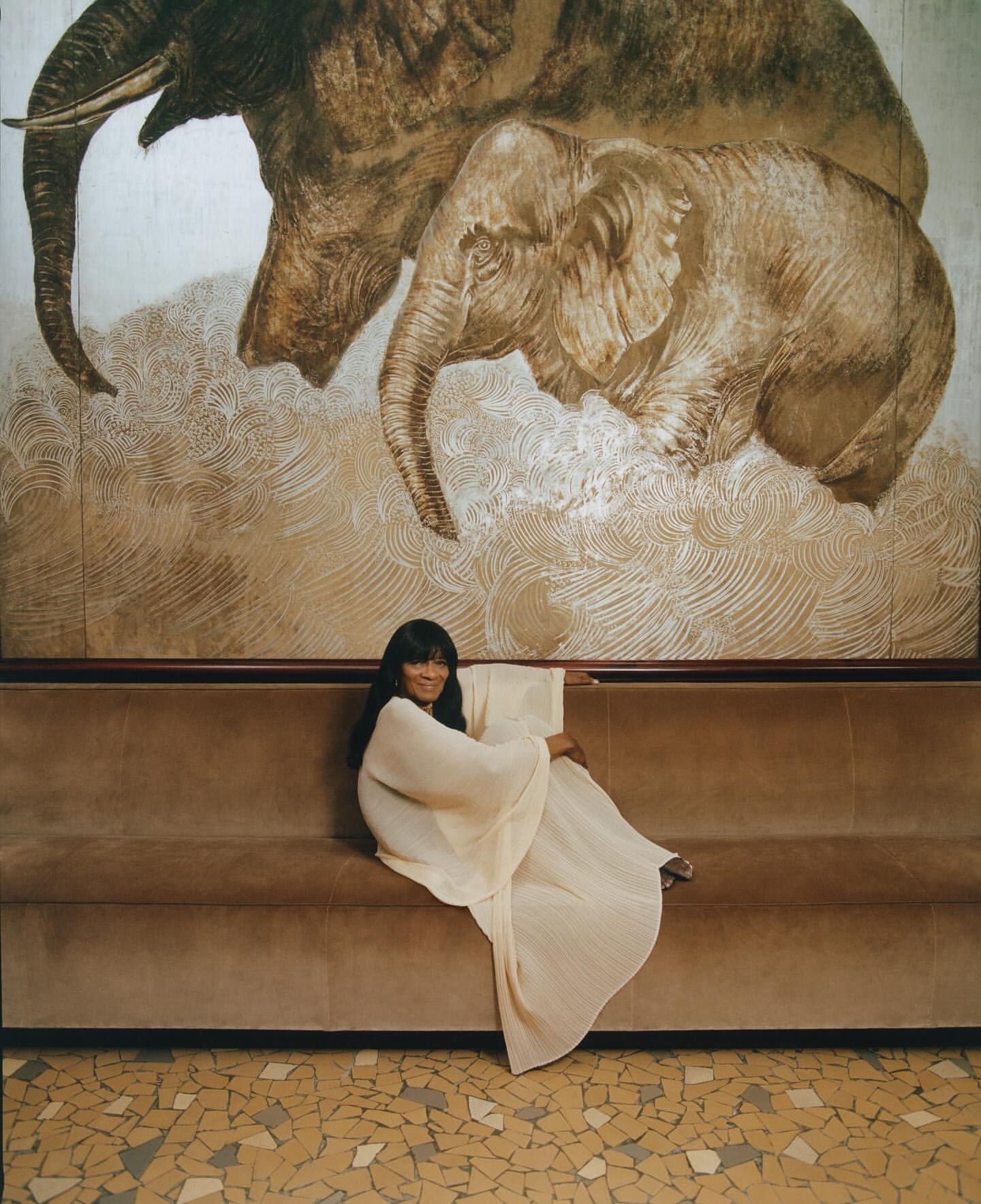Woman with long dark hair sits on a brown couch in front of a wall with elephant art, wearing a long white dress, inside a room.