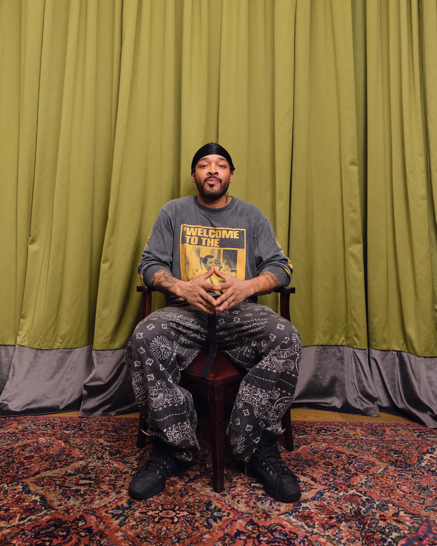Portrait of an adult man sitting in a chair against a green curtain backdrop, wearing a long-sleeve shirt and paisley print pants.