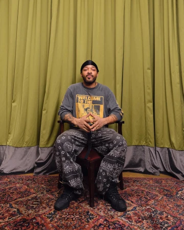 Portrait of an adult man sitting in a chair against a green curtain backdrop, wearing a long-sleeve shirt and paisley print pants.