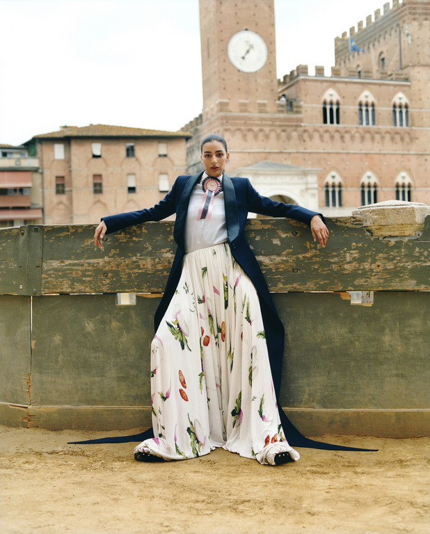 Woman in stylish floral skirt and dark blue blazer poses against an old wooden barrier, with a brick tower in the background.