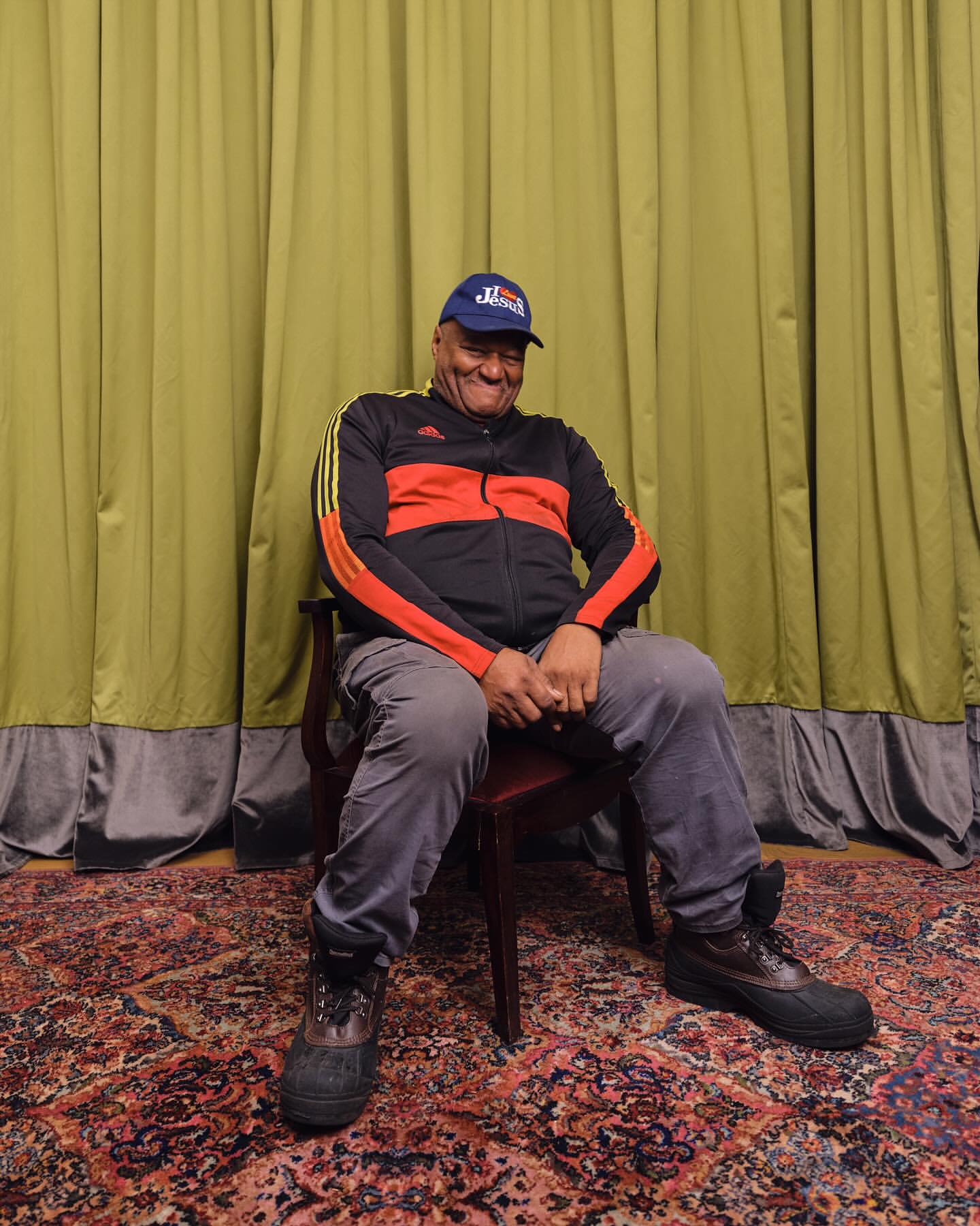 An adult man in a tracksuit and cap sits on a red chair indoors. A patterned rug is on the floor, and a green curtain is behind him.