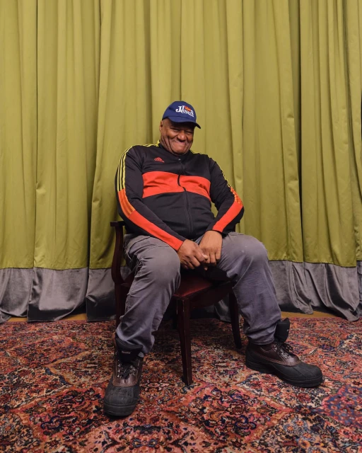 An adult man in a tracksuit and cap sits on a red chair indoors. A patterned rug is on the floor, and a green curtain is behind him.