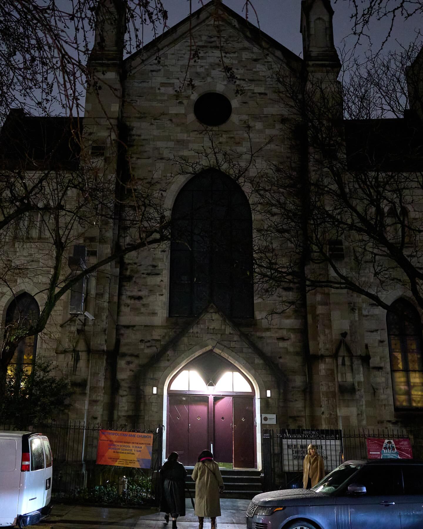 Exterior view of a stone church at night, with two people standing at the entrance and a parked car and van on the street.