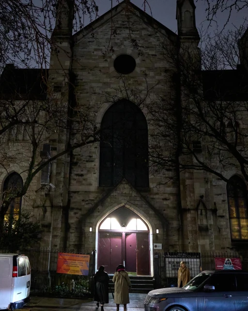 Exterior view of a stone church at night, with two people standing at the entrance and a parked car and van on the street.