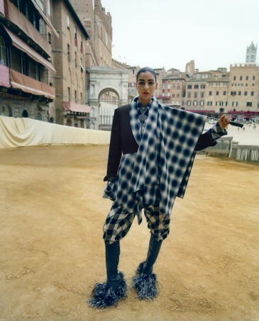 A woman in patterned clothing stands in an Italian piazza, wearing a jacket, scarf, bloomers and fuzzy heeled boots with historic buildings.