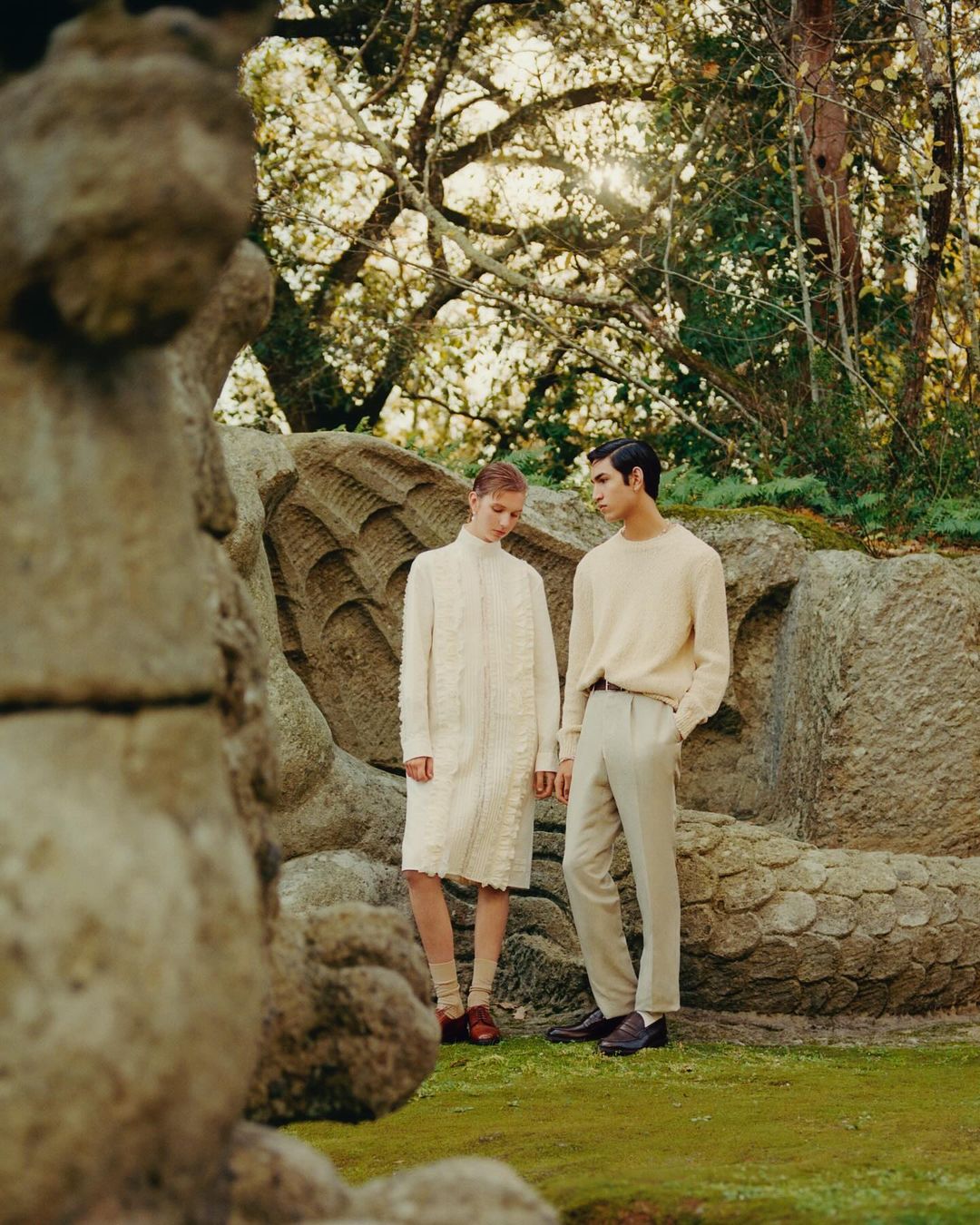 Two adults in cream-colored clothing stand on the grass near a stone structure in a forest landscape during the daytime.