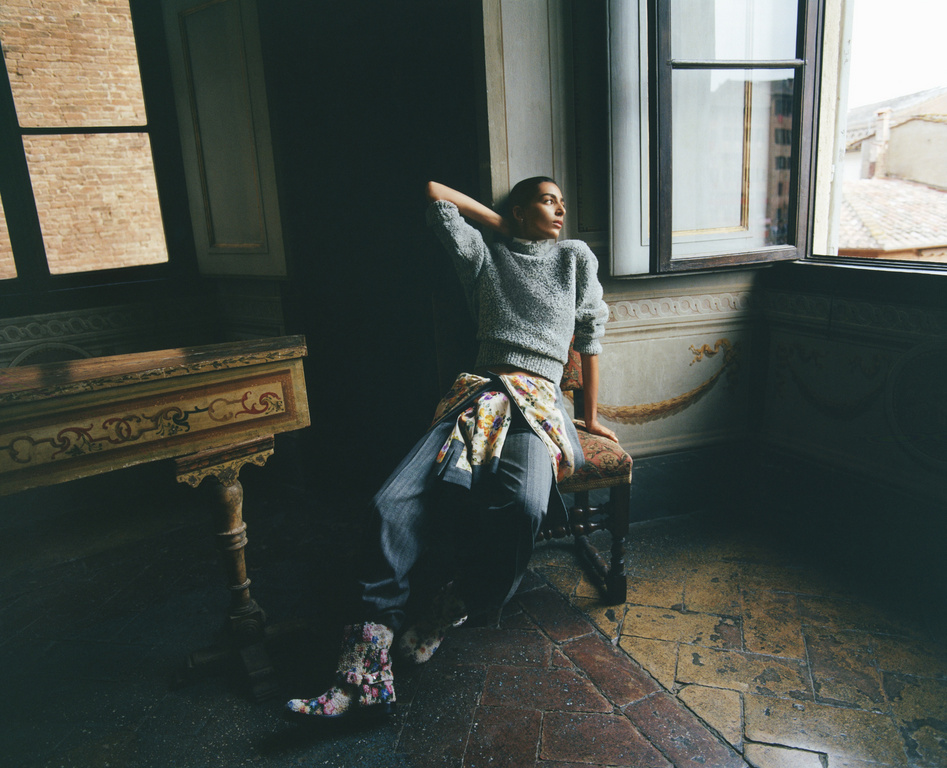 An adult woman in a grey sweater and blue jeans sits on a chair near a window inside a vintage style room with painted walls.
