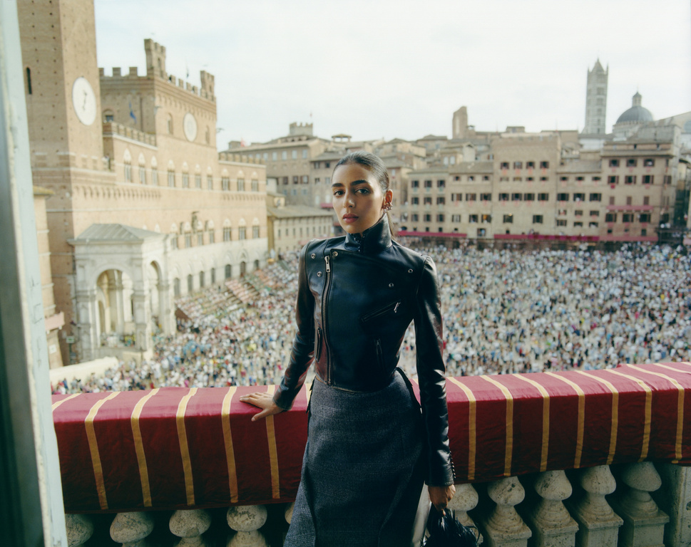 Woman in black leather jacket stands on a balcony overlooking a large crowd in an urban square with historic buildings and towers.