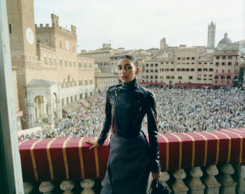Woman in black leather jacket stands on a balcony overlooking a large crowd in an urban square with historic buildings and towers.