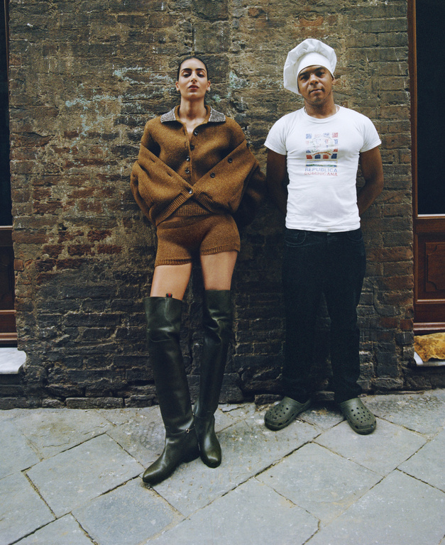 A woman in a brown knit outfit and black boots poses next to a man in a chef's hat against a textured brick wall on a tiled floor.