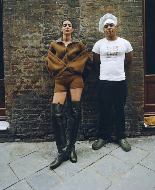 A woman in a brown knit outfit and black boots poses next to a man in a chef's hat against a textured brick wall on a tiled floor.