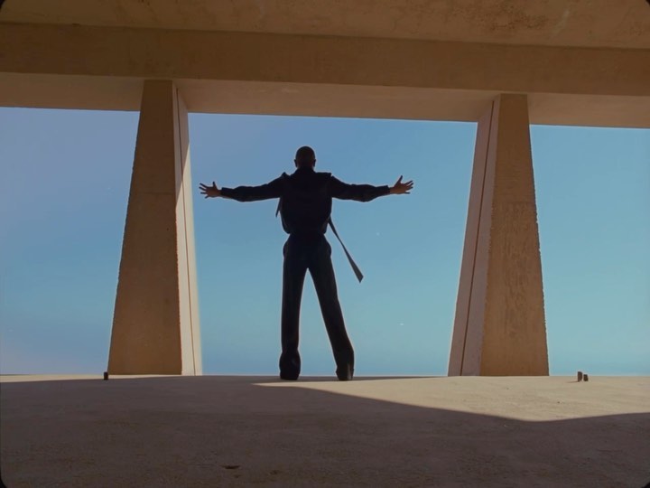 Silhouette of a man with arms outstretched, standing against a light blue sky, framed by geometric architectural elements.