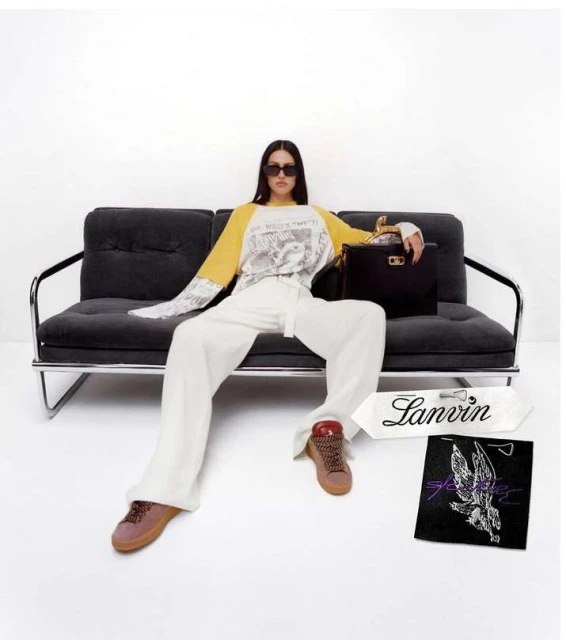 A woman in white pants and a color-blocked shirt poses on a black couch with a black handbag and Lanvin-branded shopping bag.