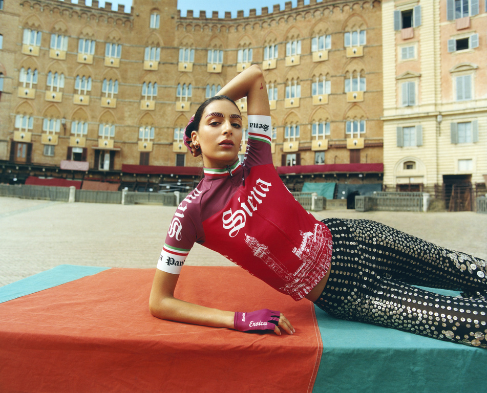 Woman in cycling jersey poses on orange and teal surface with Siena text, in front of building with arched windows on piazza.