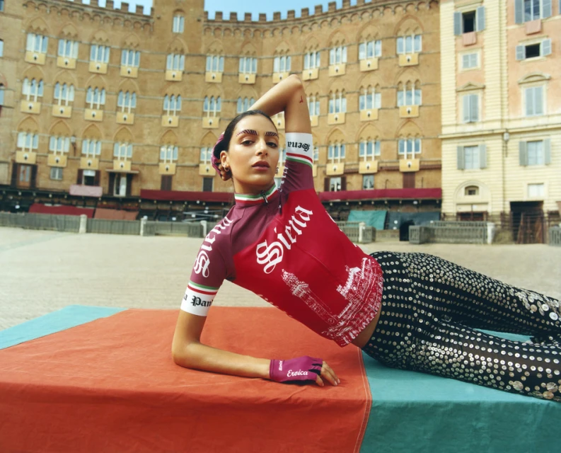 Woman in cycling jersey poses on orange and teal surface with Siena text, in front of building with arched windows on piazza.