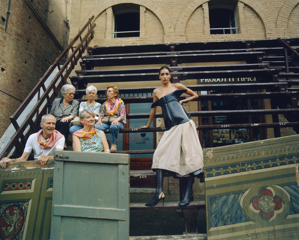 Fashion photo with a model in a corset and skirt posing on a staircase with elderly people in the background. The shot is outdoors.