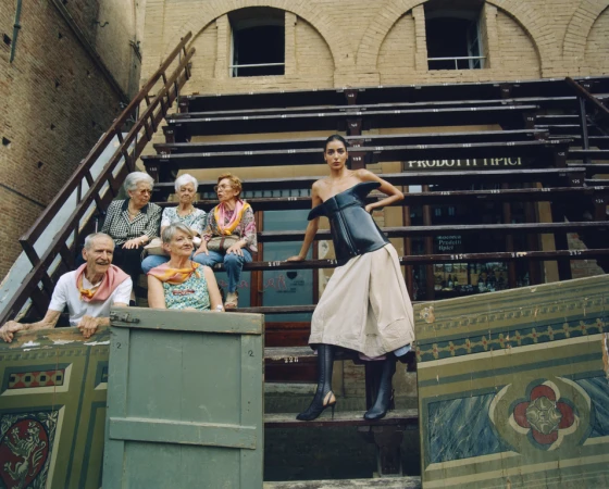 Fashion photo with a model in a corset and skirt posing on a staircase with elderly people in the background. The shot is outdoors.