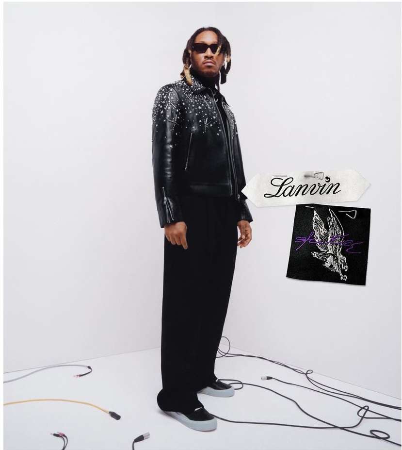 Portrait of a Black male model in a studded leather jacket and black pants, posing in a white photo studio next to brand labels.