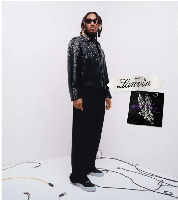Portrait of a Black male model in a studded leather jacket and black pants, posing in a white photo studio next to brand labels.
