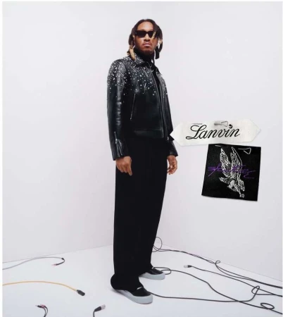Portrait of a Black male model in a studded leather jacket and black pants, posing in a white photo studio next to brand labels.