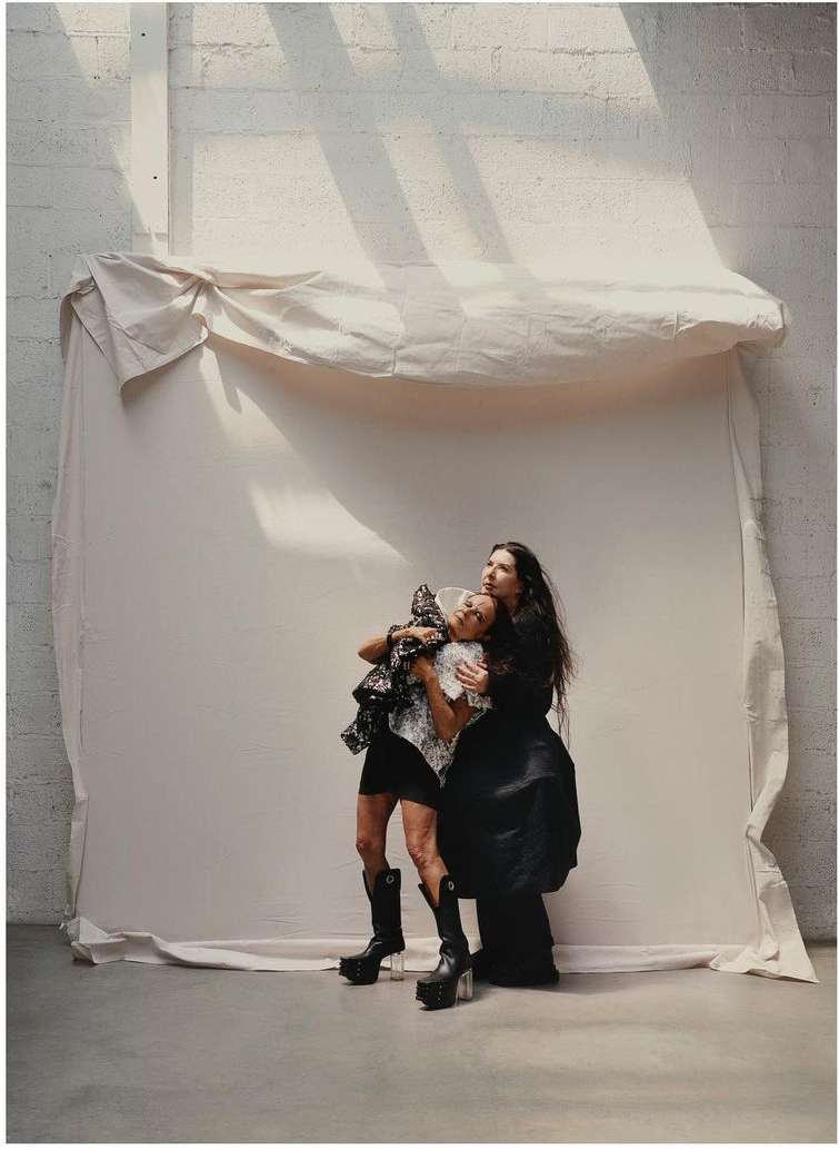 Two women in dark clothing are close together in a photo studio with a canvas backdrop. One woman wears platform boots and hugs the other.