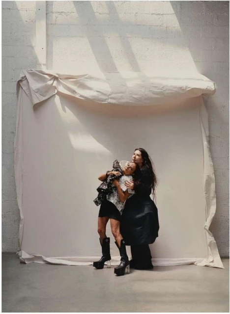 Two women in dark clothing are close together in a photo studio with a canvas backdrop. One woman wears platform boots and hugs the other.