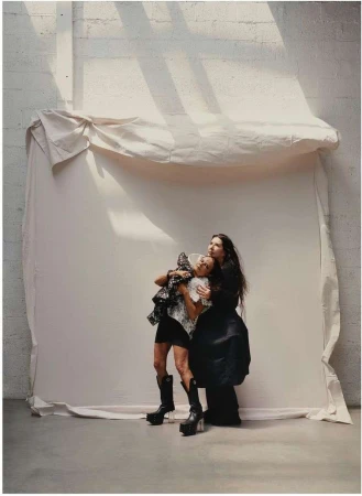 Two women in dark clothing are close together in a photo studio with a canvas backdrop. One woman wears platform boots and hugs the other.