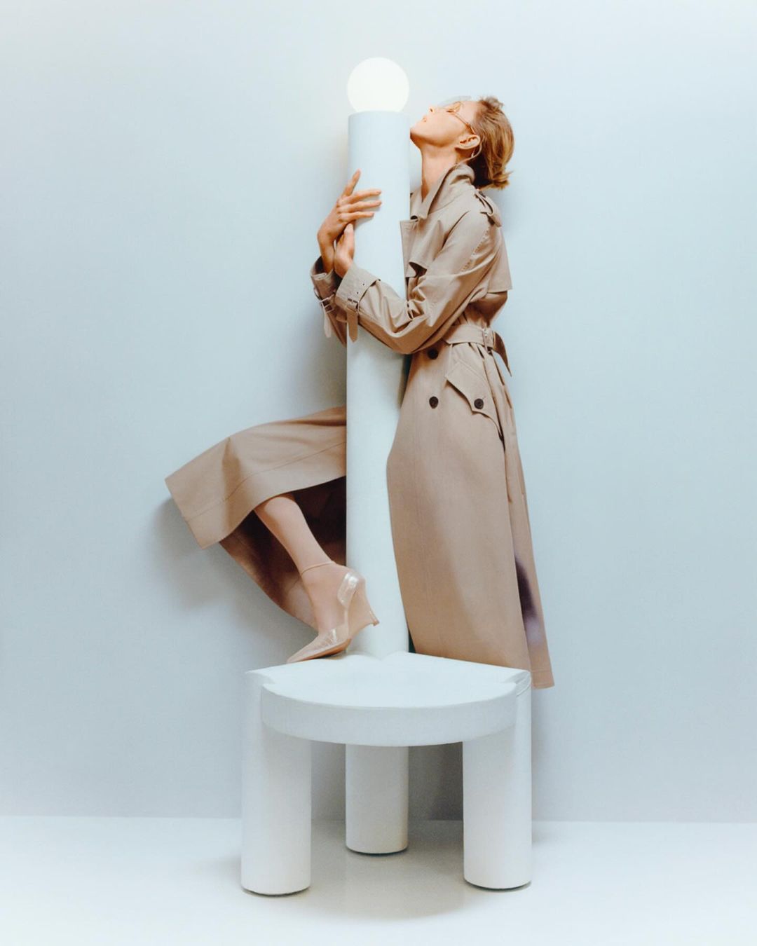Woman in a tan trench coat posing on a white platform with a white column light, set against a pale blue-grey background.