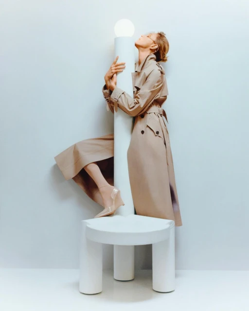 Woman in a tan trench coat posing on a white platform with a white column light, set against a pale blue-grey background.
