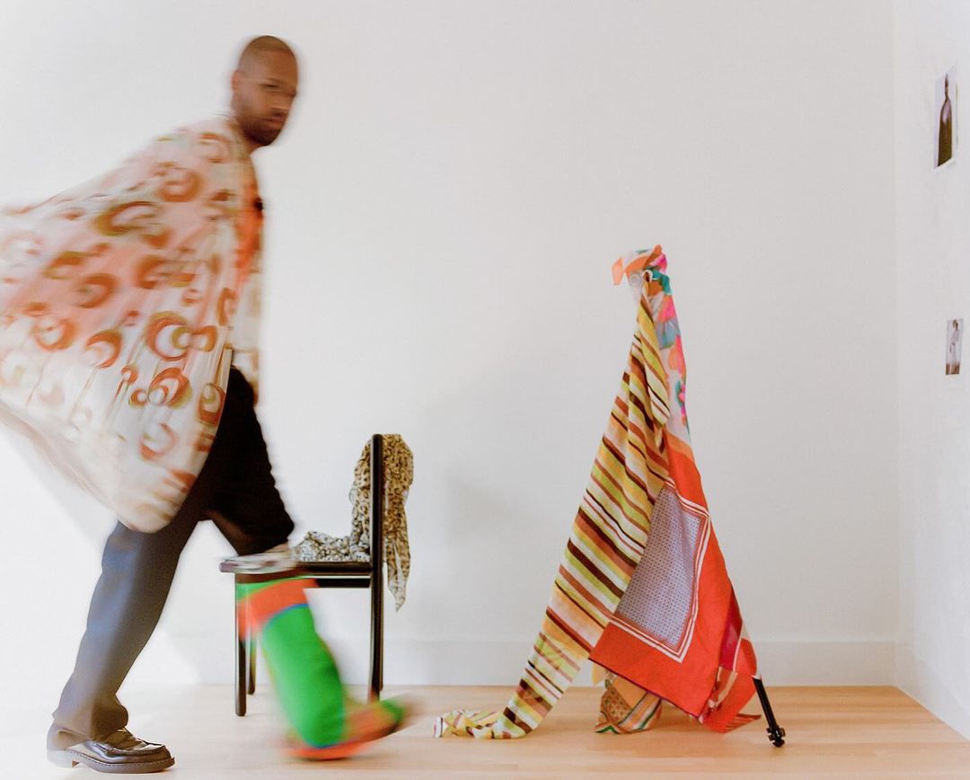 Blurred figure in patterned robe walking by chair with draped fabrics, set against a stark white wall and wooden floor.