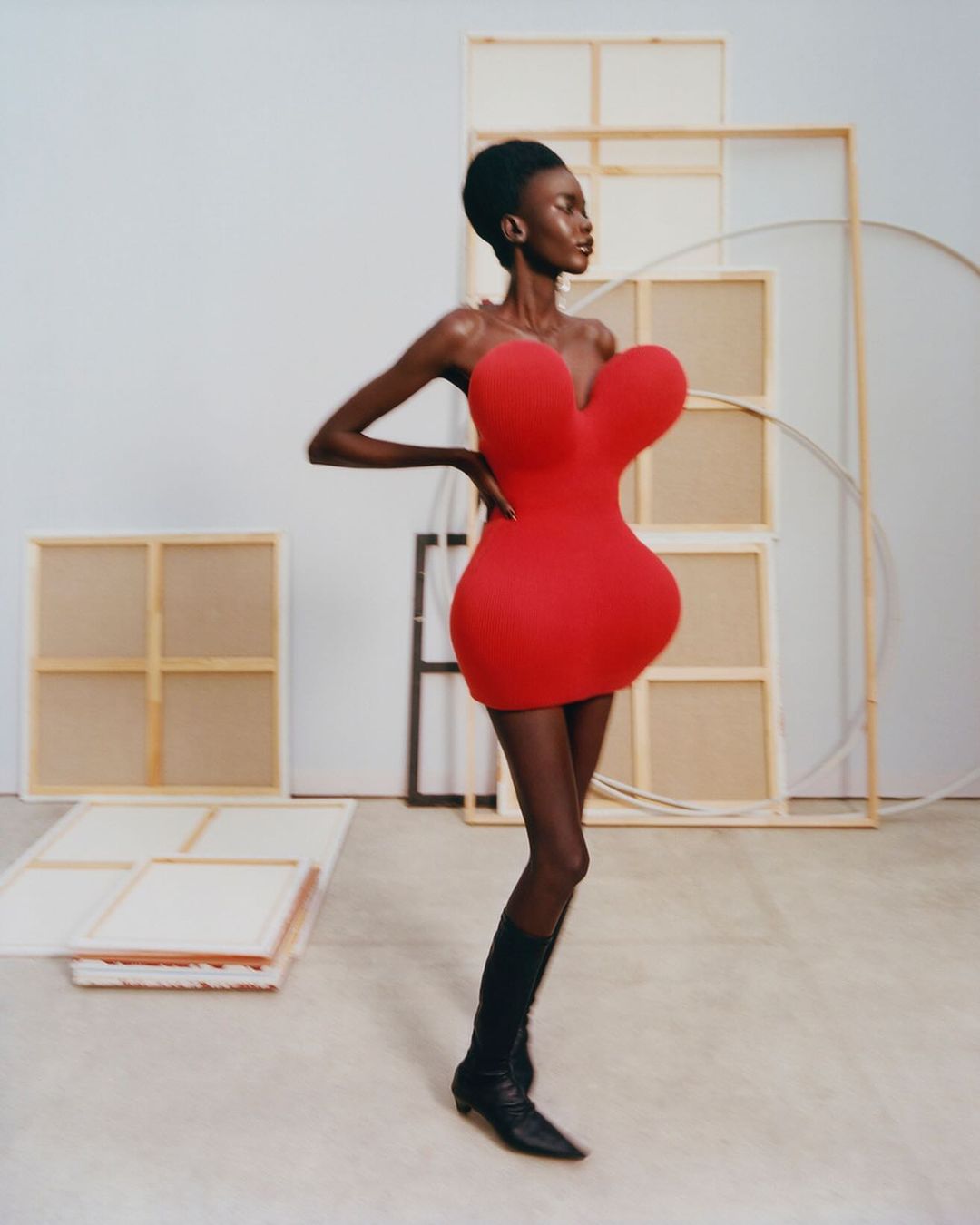 An African American female model poses in a red mini dress and black boots against a plain white backdrop with art supplies.