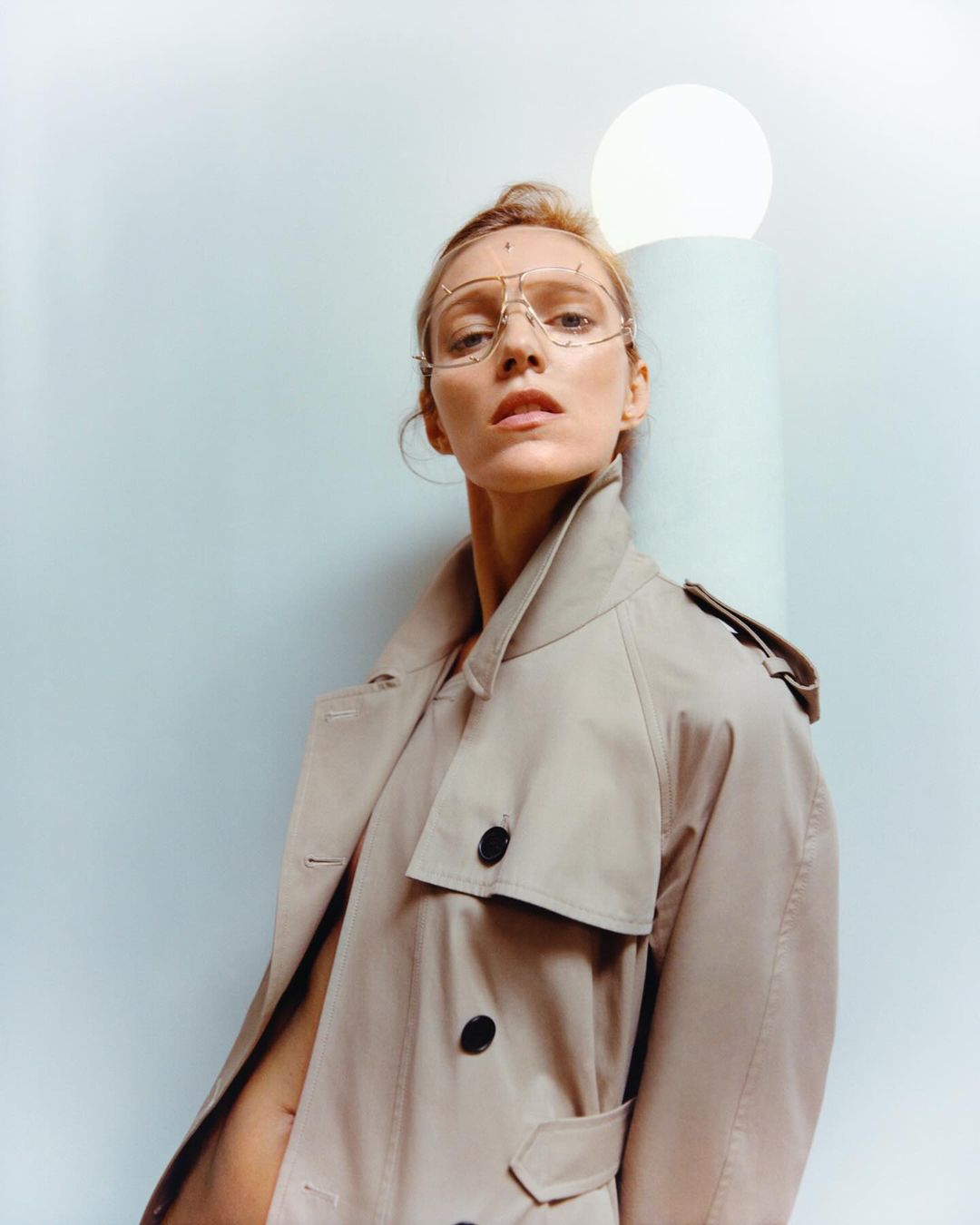 Portrait of a blonde adult woman wearing a trench coat and stylized glasses against a blue and white backdrop with a light.