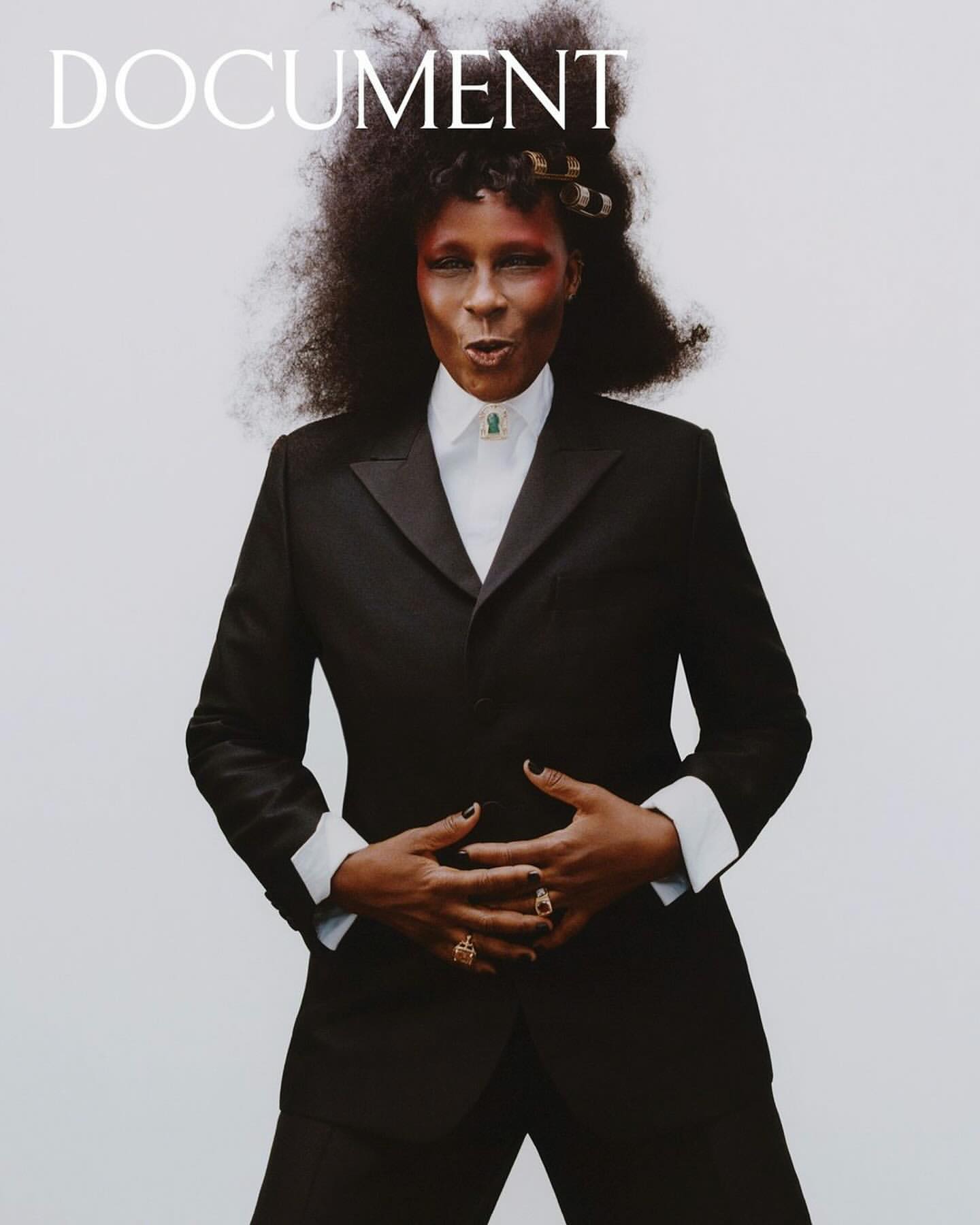 Elegant portrait of a Black woman in a black suit with afro hair, adorned with gold hair clips. She is wearing rings and a necklace.