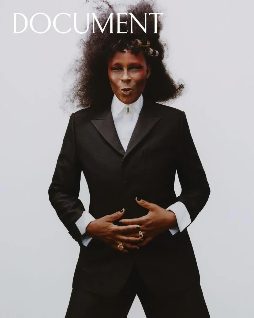 Elegant portrait of a Black woman in a black suit with afro hair, adorned with gold hair clips. She is wearing rings and a necklace.