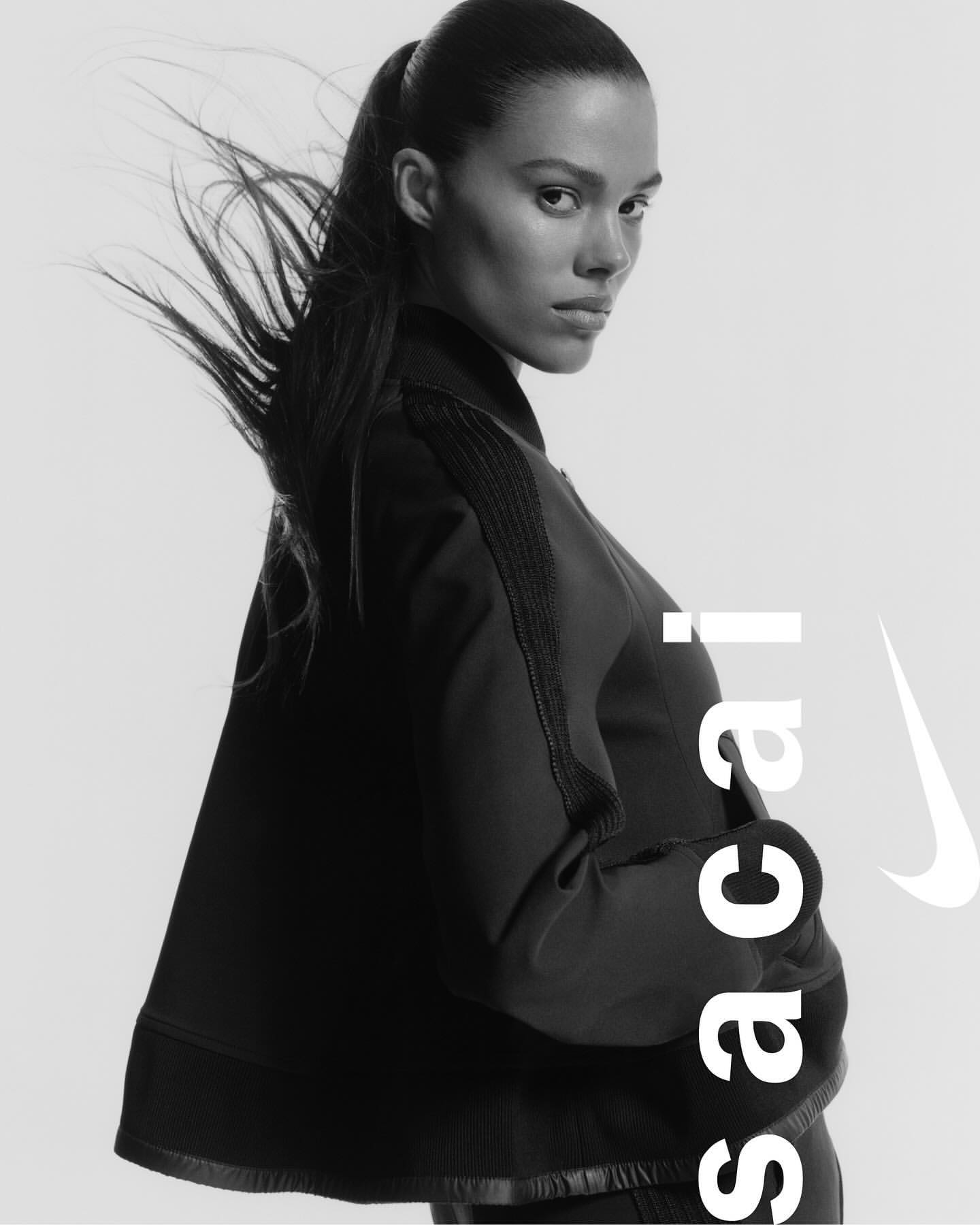 Monochrome portrait of an adult female model wearing a black jacket by Nike x Sacai against a light backdrop, showcasing fashion and style.