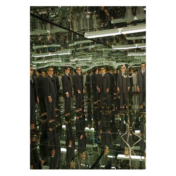 Multiple adult men in dark suits and sunglasses stand in a reflective space creating a mirrored effect of the group and bright overhead lights.