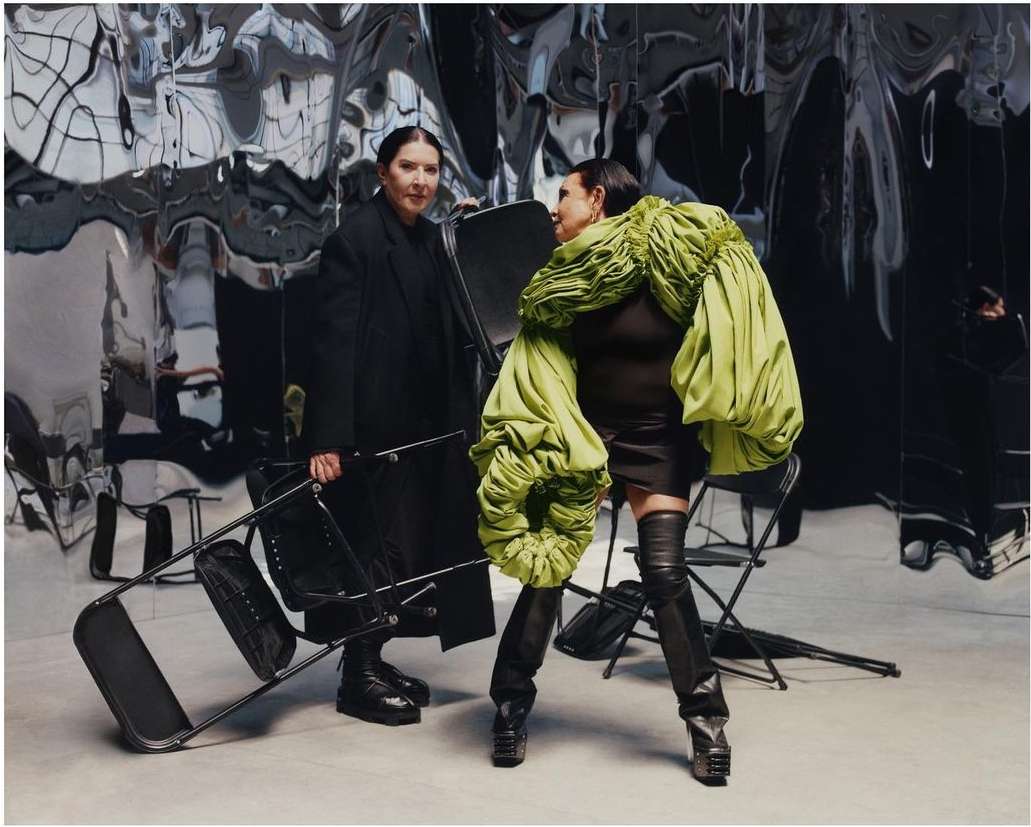 Two women pose indoors against a reflective backdrop; one holds a metal chair, the other wears a green sculpted garment and black thigh-high boots.