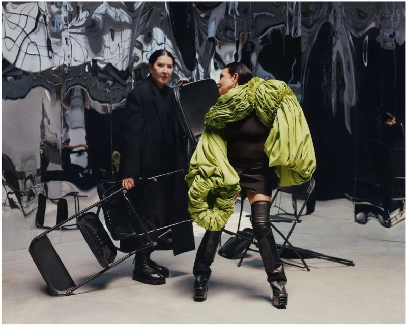 Two women pose indoors against a reflective backdrop; one holds a metal chair, the other wears a green sculpted garment and black thigh-high boots.